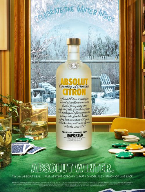 Absolut Citron vodka bottle stands on a poker table with a snowy winter scene visible through a window behind it.
