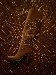 Brown suede knee-high heeled boot with gold studded paisley design and fringe against a patterned studded background.