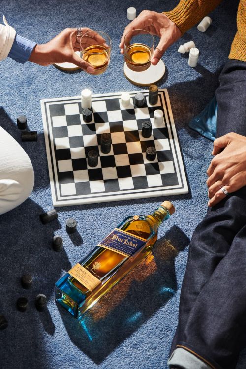 Two adults enjoying whiskey while playing checkers on a checkered board atop a soft blue carpet, featuring a Blue Label bottle.