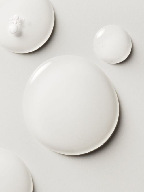 Elevated view of semi-translucent white liquid spheres of different sizes on a pale grey surface, creating an abstract, minimalist composition.