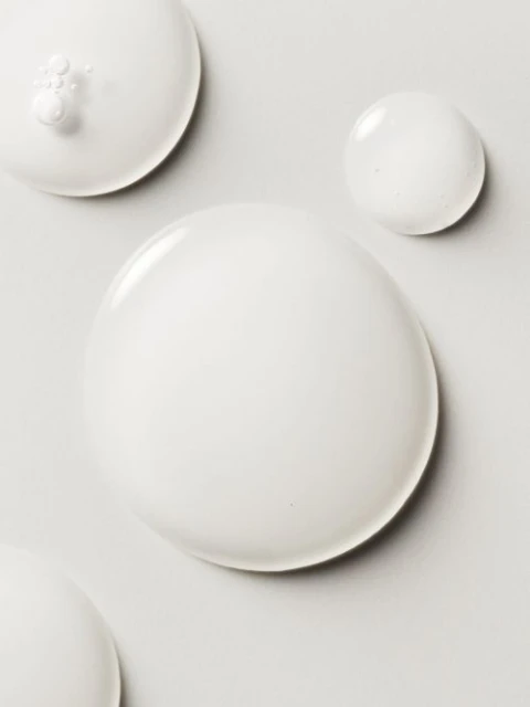 Elevated view of semi-translucent white liquid spheres of different sizes on a pale grey surface, creating an abstract, minimalist composition.
