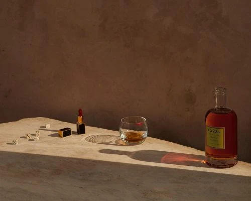Still life shot of a liquor bottle, glass with liquid, dice, and lipstick on a beige surface in a photo studio with neutral background.