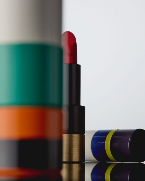 Close-up product shot of red lipstick, open and standing, with striped packaging details on a reflective black surface against white.