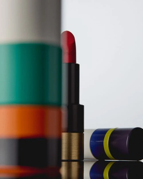 Close-up product shot of red lipstick, open and standing, with striped packaging details on a reflective black surface against white.