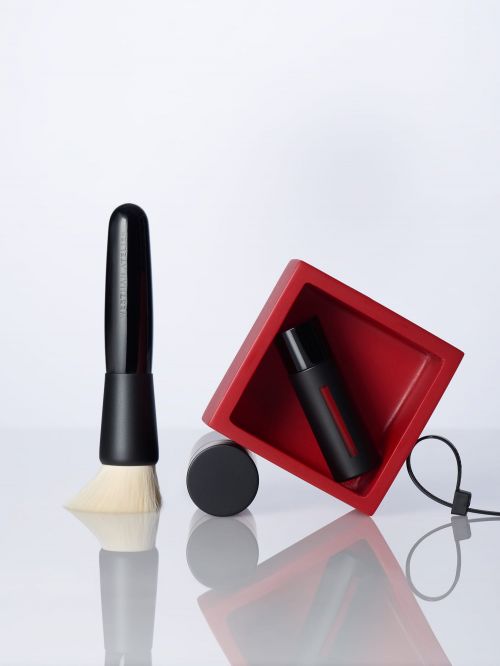 Still life shot of black makeup brush, makeup container, and red display case against a white backdrop, with reflections on the surface.