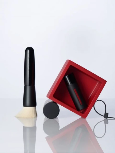 Still life shot of black makeup brush, makeup container, and red display case against a white backdrop, with reflections on the surface.