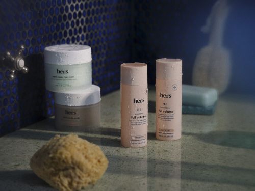 Still life of Hers brand hair-care products in pastel pink and neutral tones, set against a blue tiled backdrop with a natural sponge.