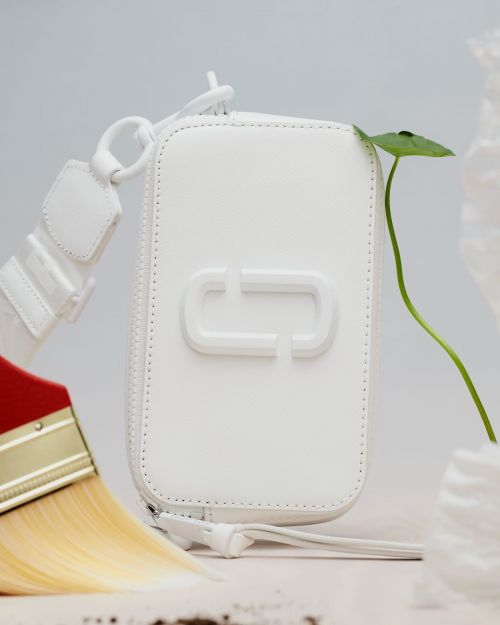 White leather purse with the brand logo on a white surface with a green sprout, a paintbrush, and decorative white elements.