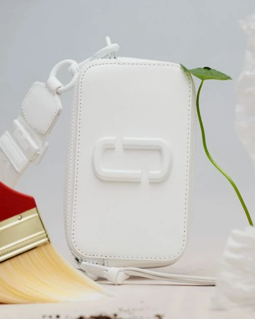 White leather purse with the brand logo on a white surface with a green sprout, a paintbrush, and decorative white elements.