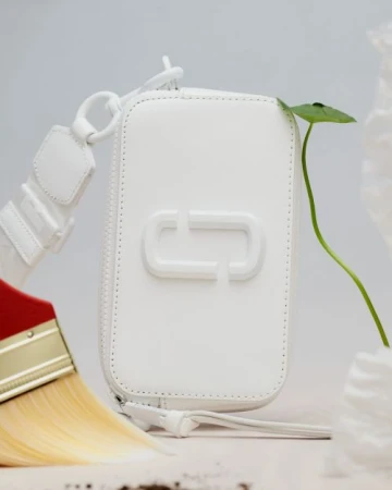 White leather purse with the brand logo on a white surface with a green sprout, a paintbrush, and decorative white elements.