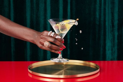 Woman's dark skin arm holding a martini glass with lemon garnish, adorned with rings, on a gold tray with red table and green curtain.