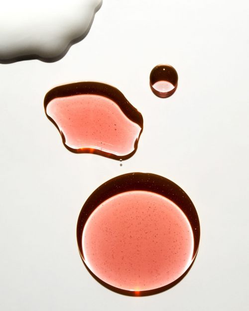 High-angle, close-up shot of cosmetic liquid blobs in varying sizes and hues of reddish-brown and white on a white background.