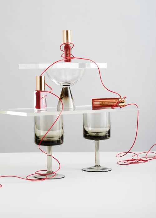 Still life of three lip glosses and geometric acrylic stands arranged on a white surface with red thread connecting the objects.