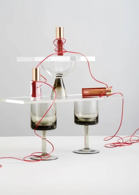 Still life of three lip glosses and geometric acrylic stands arranged on a white surface with red thread connecting the objects.