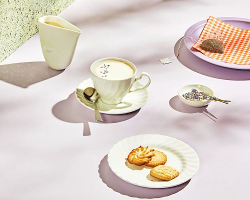 Overhead shot of tea service featuring teacup, saucer, milk pitcher, cookies, plate, and lavender on a pastel background with soft shadows.