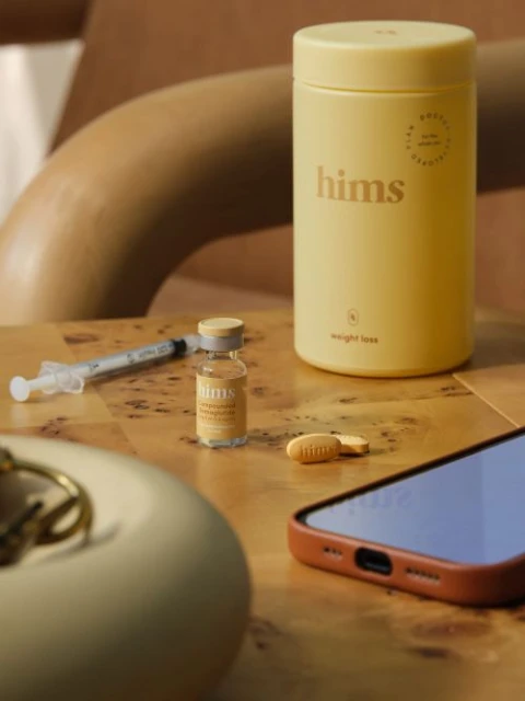 Hims brand weight loss products on a wooden surface with a cell phone, including a bottle of medication and a syringe.