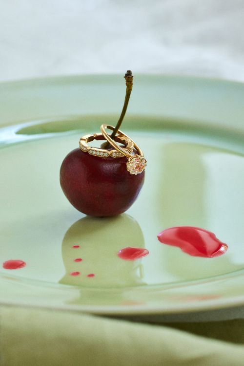 Close-up of gold diamond rings resting on a ripe red cherry atop a pale green glazed ceramic plate with fruit juice droplets