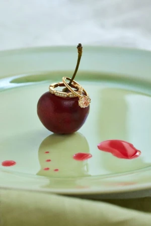 Close-up of gold diamond rings resting on a ripe red cherry atop a pale green glazed ceramic plate with fruit juice droplets