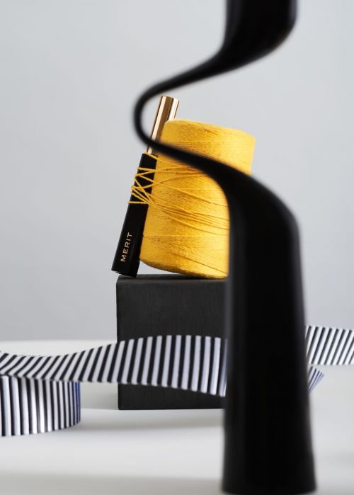 Still life of a spool of yellow thread with a black and gold makeup applicator, resting on a black cube, accented by black and white ribbon.