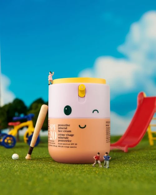 Miniature playground scene featuring a sunscreen bottle designed with a smiling face, baseball, and tiny figures against a blue sky backdrop.