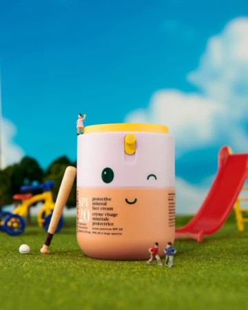 Miniature playground scene featuring a sunscreen bottle designed with a smiling face, baseball, and tiny figures against a blue sky backdrop.