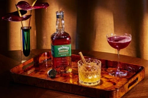 Bottle of Jack Daniel's Bonded Rye whiskey, cocktails, and a vase with flowers arranged on a wooden serving tray.