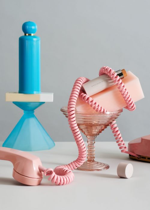 Still life photo of a vintage pink telephone with colorful geometric objects and cosmetic container against a light gray background.