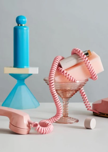 Still life photo of a vintage pink telephone with colorful geometric objects and cosmetic container against a light gray background.
