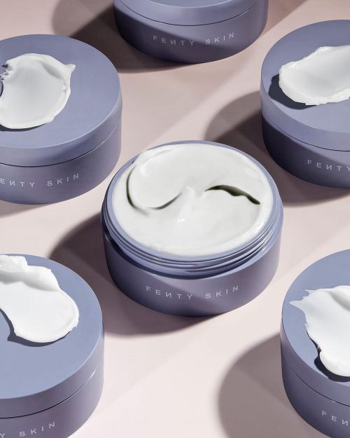 Several purple Fenty Skin jars are arranged on a pink surface, some with a white cream resting on top of the closed container.