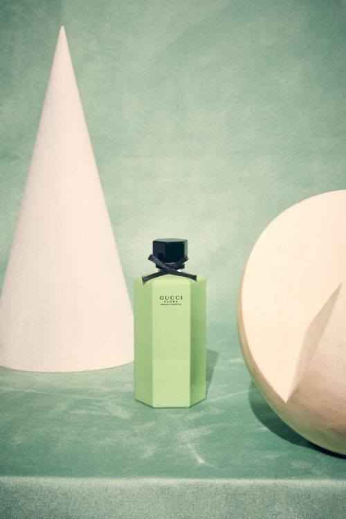 Product shot of light-green Gucci Flora Gorgeous Magnolia perfume bottle with a black cap, against a geometric set backdrop.