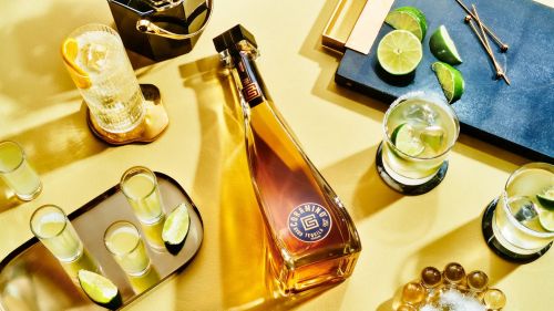 Overhead shot of a bottle of Coramino tequila with shot glasses and cocktails, garnished with lime and orange on a yellow surface.