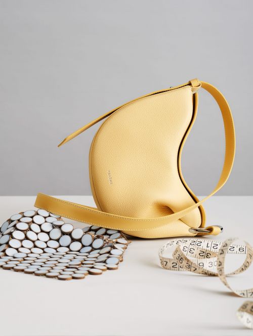 Yellow leather handbag with a unique, crescent-like shape, alongside a white and brown tile piece and measuring tape on a table.