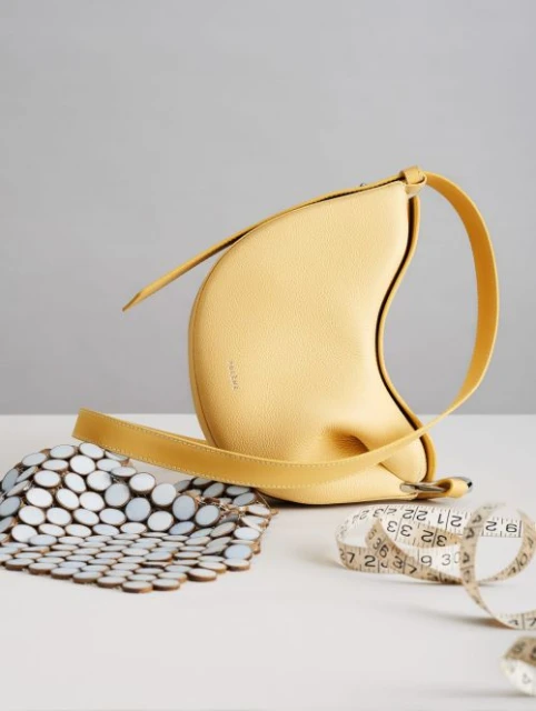 Yellow leather handbag with a unique, crescent-like shape, alongside a white and brown tile piece and measuring tape on a table.