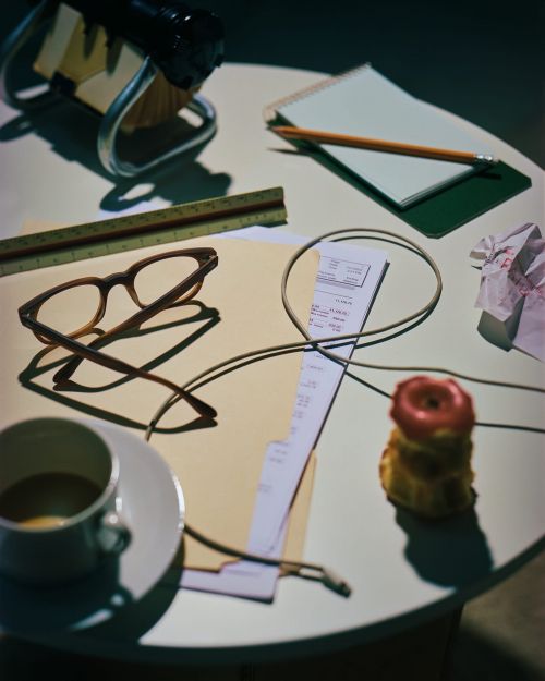 Overhead shot of a desk with an apple core, coffee cup, glasses, stationery, ruler, crumpled paper, and charging cable.