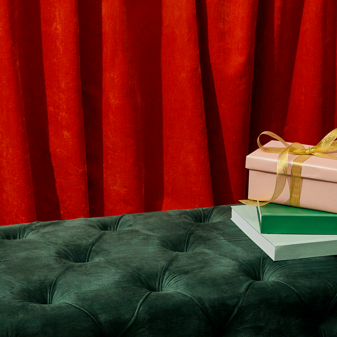 Presents with gold ribbons stacked on books atop a green velvet tufted bench in front of a red velvet curtain backdrop.