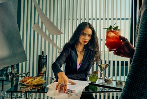 Elegant woman in black blazer works at modern desk, drinks nearby, vertical blinds in background, elevated view.