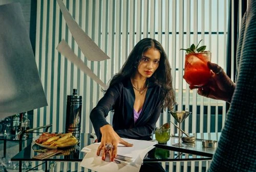 Elegant woman in black blazer works at modern desk, drinks nearby, vertical blinds in background, elevated view.
