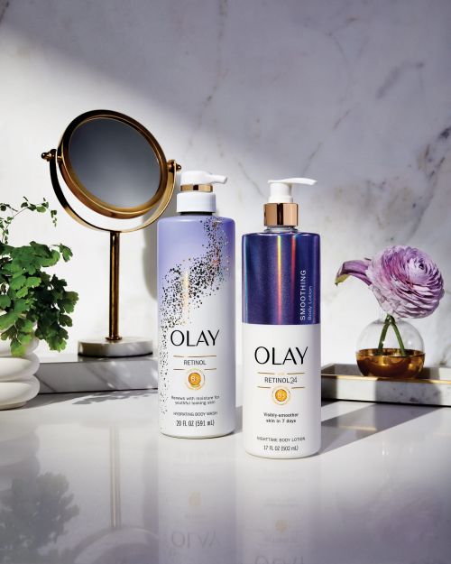 Two bottles of Olay Retinol body wash and body lotion are displayed on a reflective surface next to a flower and a round mirror.