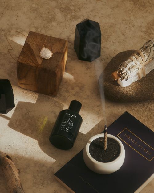 Still life arrangement featuring sage bundle, incense stick in holder, and various crystals and stones, set against a neutral surface.