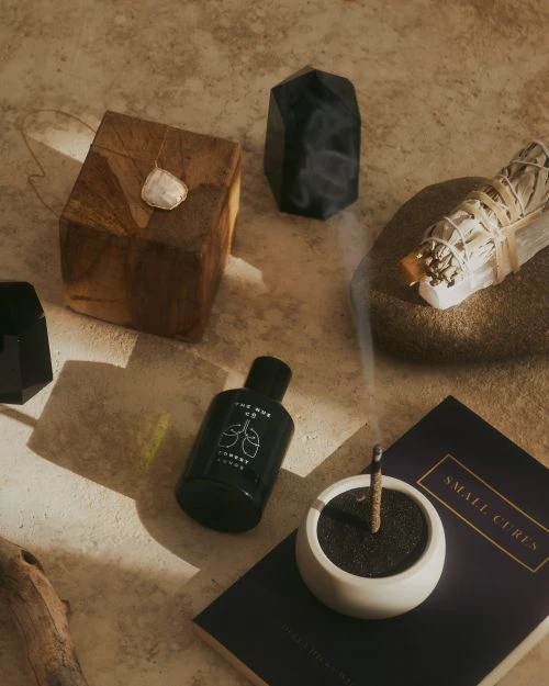 Still life arrangement featuring sage bundle, incense stick in holder, and various crystals and stones, set against a neutral surface.