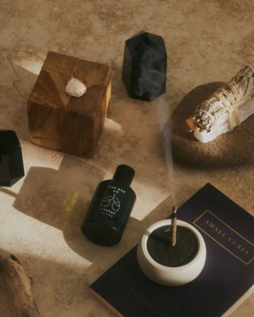 Still life arrangement featuring sage bundle, incense stick in holder, and various crystals and stones, set against a neutral surface.