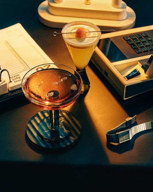 Two cocktail glasses sit on a dark surface near a day planner, calculator, and watch under studio lighting, creating an intimate still life.