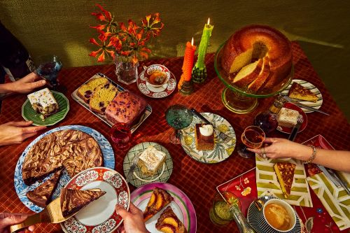 Overhead shot of a table laden with cakes, slices, and teacups, surrounded by hands, lit by candles, set against a textured green wall.