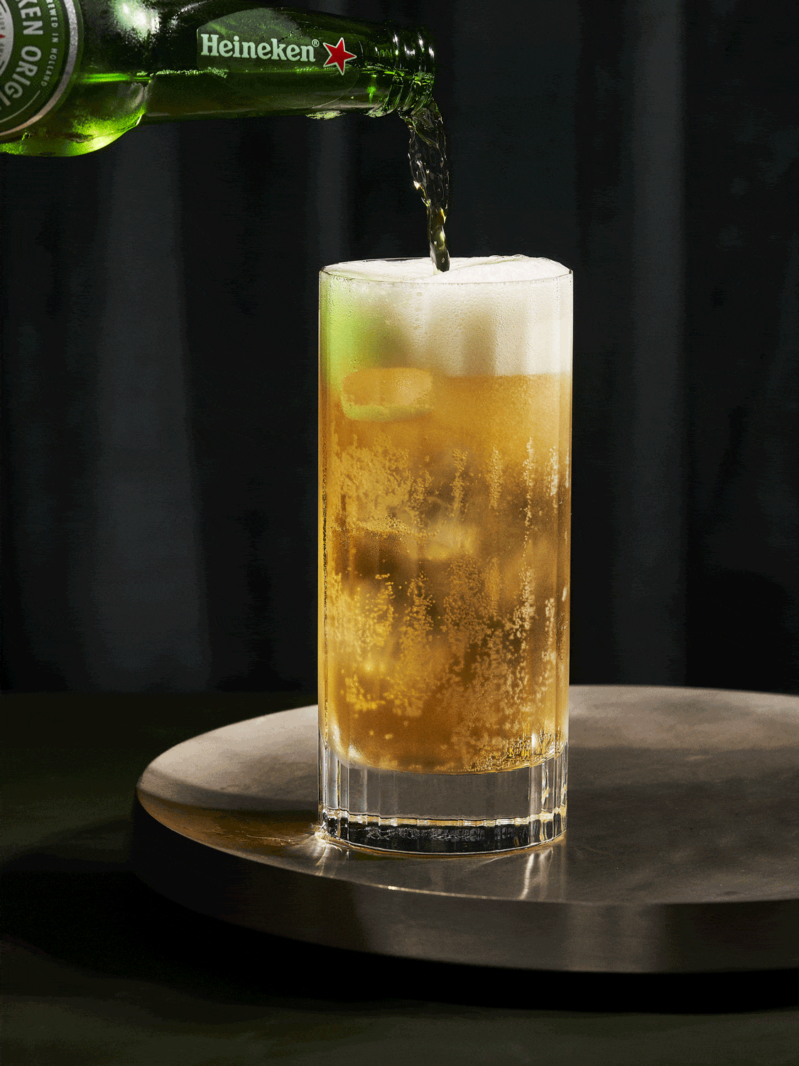 Heineken beer bottle pours golden liquid into a tall, foamy glass on a silver tray against a dark backdrop.