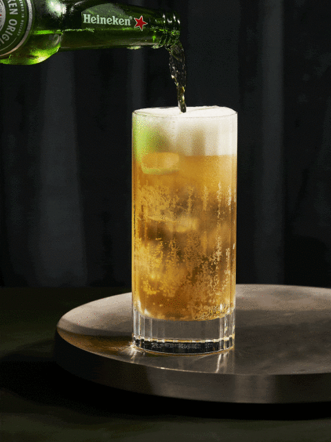 Heineken beer bottle pours golden liquid into a tall, foamy glass on a silver tray against a dark backdrop.