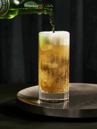 Heineken beer bottle pours golden liquid into a tall, foamy glass on a silver tray against a dark backdrop.
