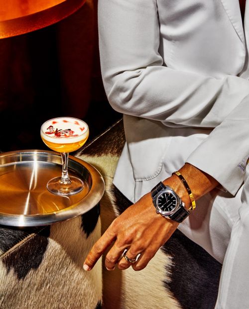 Cocktail on a silver tray with an adult wearing a white suit and a watch, seated on a brown and white animal print bench seat.
