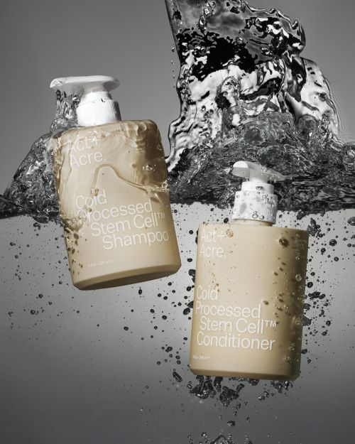 Two bottles of Act+Acre Cold Processed Stem Cell shampoo and conditioner partially submerged in water with splashing droplets.