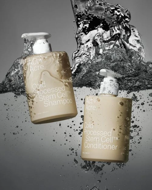 Two bottles of Act+Acre Cold Processed Stem Cell shampoo and conditioner partially submerged in water with splashing droplets.