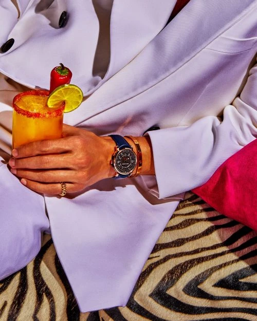Person in a white suit holds a cocktail garnished with chili pepper and lime slice. Watch and jewelry visible. Zebra print background.